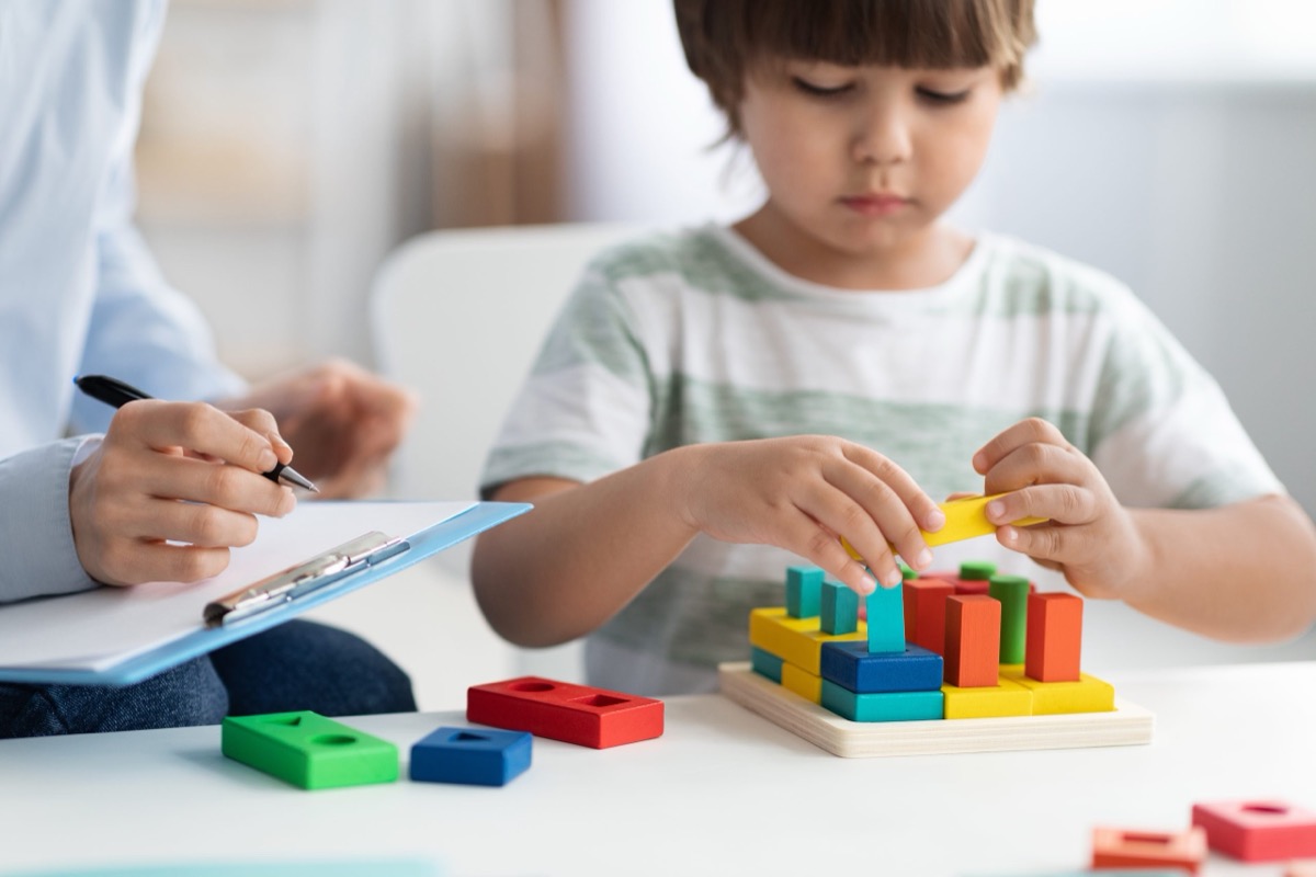 A young child builds with colorful blocks while a therapist takes notes during a play therapy session